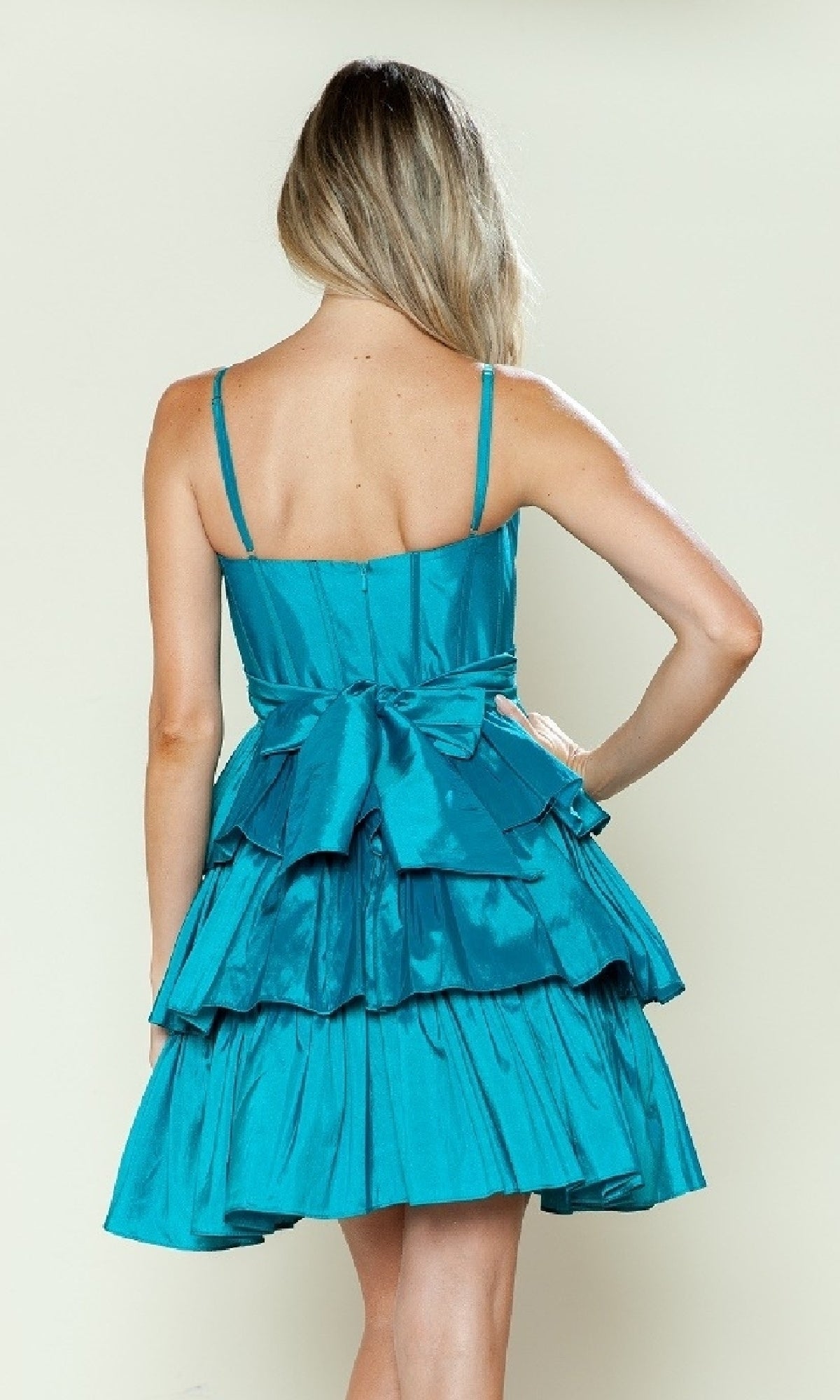 A woman stands with her back to the camera, modeling the Poly USA 9590 Short Homecoming Dress in teal—a sleeveless fit-and-flare style with a tiered skirt and statement waist bow.
